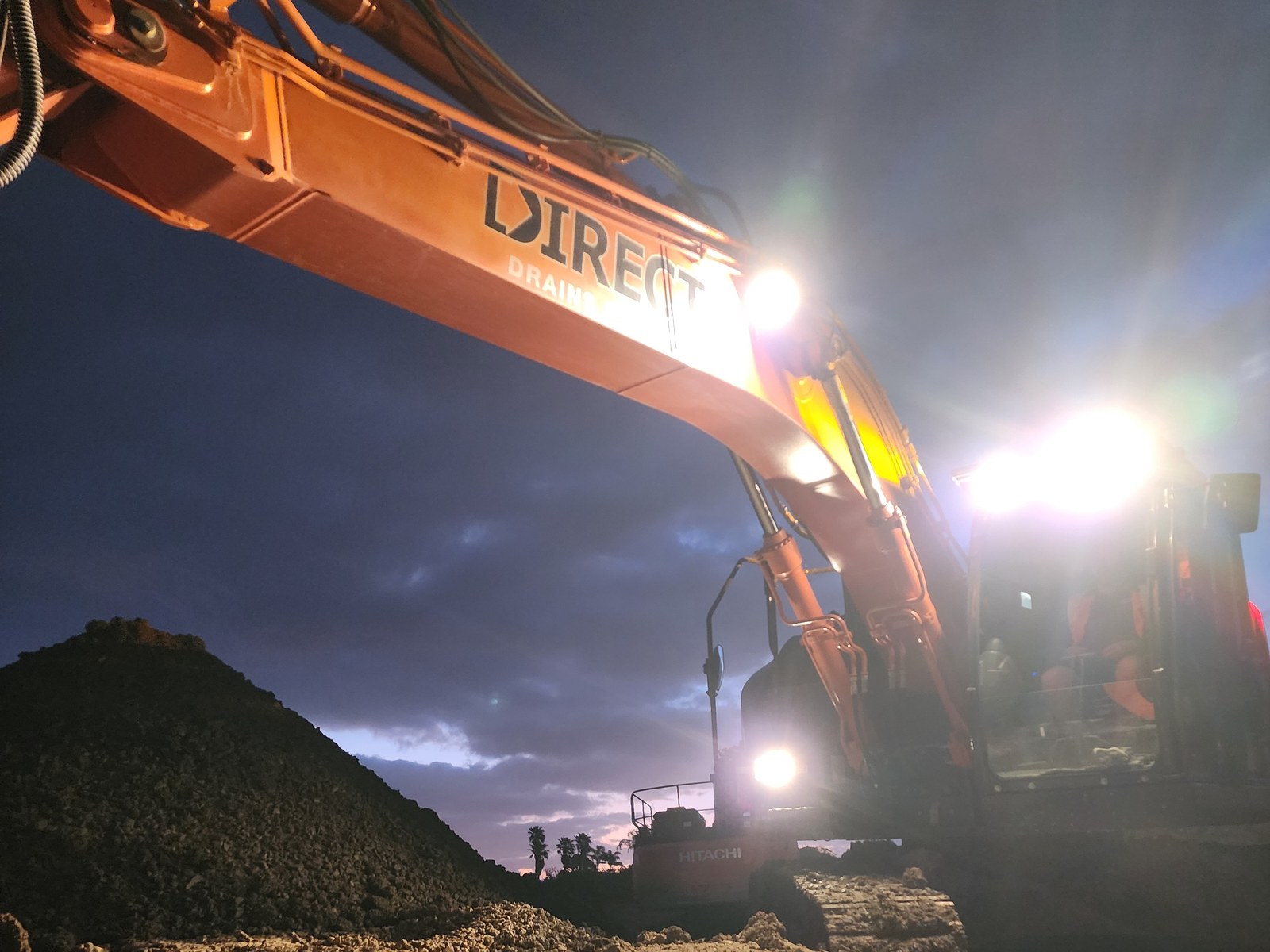 A Direct Drains excavator working an open drainage trench under lights at dusk.