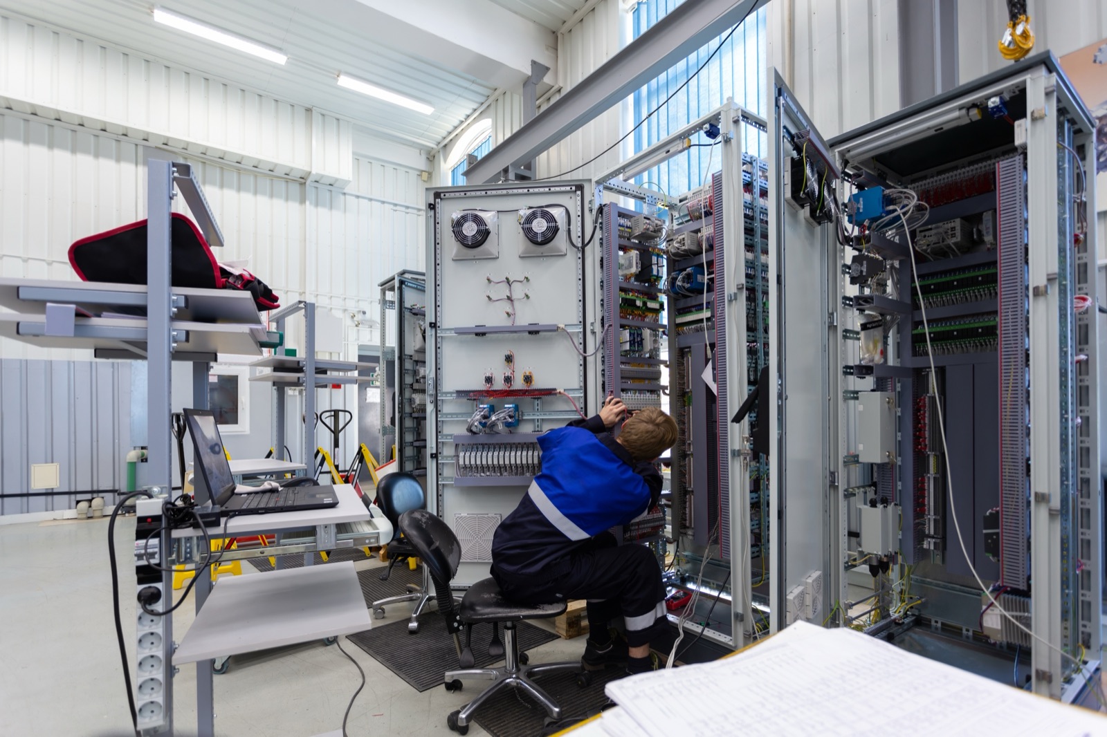 A technician wiring switchboard cabinets on the workshop floor, test gear and drawings alongside